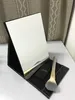 A makeup mirror and brush are displayed on a white surface with a patterned curtain in the background. Produktbild-Vorschau