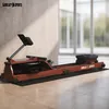 A rowing machine is placed on a mat in front of a wall and large windows. Produktbild-Vorschau