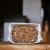 A loaf of bread with seeds and grains, presented in a paper liner within a box, placed on a wooden surface. Produktbild-Vorschau