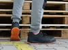 A person wearing black safety shoes with orange soles stands in front of wooden pallets. Produktbild-Vorschau