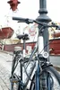 A bicycle is locked to a lamppost in a harbor area with boats and buildings in the background. Produktbild-Vorschau