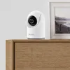 A white Reolink E Series E321 indoor camera sits on a wooden surface next to a framed picture. Náhled obrázku produktu