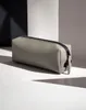 A minimalist product shot of a gray accessory case with a black zipper, placed on a white surface with dramatic shadows. Voir les images du produit
