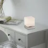 A modern cube-shaped table lamp with a frosted glass shade sits on a white dresser next to an open magazine. Anteprima dell’immagine prodotto