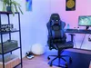 A gaming setup with a blue and black gaming chair, a desk with a computer, and shelves with gaming-related items. Produktbild-Vorschau