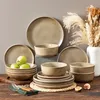A collection of stoneware dinnerware, including plates and bowls, arranged on a wooden table with some decorative elements like pampas grass and figs. Náhled obrázku produktu