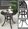 A round, dark brown metal side table with decorative elements, placed outdoors on a paved patio with plants in the background. Insets show close-ups of the table's design and construction. Podgląd zdjęcia produktu