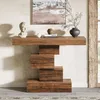 A wooden console table with a unique stacked block design, placed against a textured wall with artwork and decorative items on top. Anteprima dell’immagine prodotto