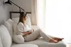 A woman sits on a sofa, holding a mug and looking out a window. Náhľad obrázku produktu