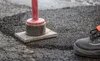 A close-up of asphalt being compacted with a tool, with a person's shoe visible on the right. Produktbild-Vorschau