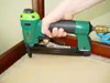 A hand is holding a green pneumatic stapler, using it to attach fabric to the corner of a wooden chair. A blue air hose is connected to the stapler. Náhľad obrázku produktu