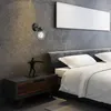 A bedroom scene with a nightstand, a wall lamp, and a bed. Produktbild-Vorschau
