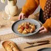 A person is placing a plate of food on a table with a runner and cutlery. Produktbild-Vorschau