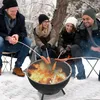 Four people are sitting around a fire pit in the snow, roasting hot dogs. Produktbild-Vorschau