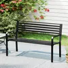 A black metal park bench sits on a paved patio in front of a white-paneled wall and lush green bushes with red flowers. Produktbild-Vorschau