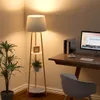 A floor lamp with shelves is placed next to a desk with a computer in a room. Podgląd zdjęcia produktu