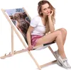 A young woman is sitting on a foldable wooden deck chair with a Scottish Cow print on a white background. Produktbild-Vorschau