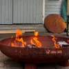 A rusty metal fire pit with flames burning, placed on a paved patio in front of a wooden building. Anteprima dell’immagine prodotto