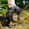 A person holding a backpack and hiking poles in a forest. Náhľad obrázku produktu