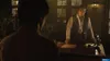 A man in a suit stands behind a desk in a dimly lit office, looking at another person. Produktbild-Vorschau