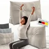 A woman is stretching while sitting on a bed with several pillows around her. Produktbild-Vorschau