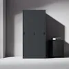 A dark gray metal filing cabinet with sliding doors is shown in a minimalist room with gray walls and a white floor. A small black cabinet with a white electronic device on top is placed to the right of the filing cabinet. Produktbild-Vorschau