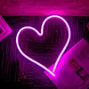 A neon heart light is placed on a dark, textured surface, with a small Eiffel Tower figurine and a folded newspaper nearby. Anteprima dell’immagine prodotto