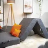 A folded mattress is set up in a living room, with two orange pillows placed on it. A floor lamp and a shelf with a picture are visible in the background. Voir les images du produit