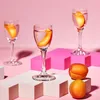 Three small glasses of clear liquid with apricot slices, arranged on white and pink geometric pedestals against a pink background. Two apricots are placed in the foreground. Produktbild-Vorschau