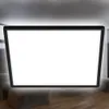 A flat, black-framed LED panel with a blank white screen is shown in a room with blurred furniture and wooden flooring. Produktbild-Vorschau