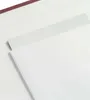 A close-up view of a white paper or cardstock, with a thin strip of lighter material running diagonally across it. A dark red textured edge is visible at the top. Produktbild-Vorschau