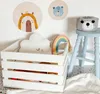 A white wooden crate filled with children's toys, including a cloud pillow and a rainbow decoration, is placed on the floor next to a light blue stool with a knitted basket on top. Two circular wooden wall decorations with animal illustrations are visible  Náhľad obrázku produktu