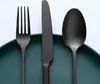 A close-up shot of a black matte cutlery set including a fork, knife, and spoon, placed on a dark green plate. Produktbild-Vorschau