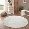 A round, fluffy white rug is placed on a wooden floor in a living room setting. The rug is in the foreground, with a ladder shelf, a basket, and a plant visible in the background. Produktbild-Vorschau
