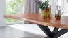 A close-up view of a wooden dining table with a live edge, set in a well-lit room with a large window and curtains in the background. A pitcher of water and two glasses are on the table, along with a potted plant. Produktbild-Vorschau