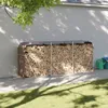 A firewood storage rack filled with logs is placed against a wall, with grass and tree shadows in the foreground. Náhled obrázku produktu