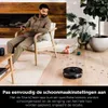 A man sits in a chair while a robot vacuum cleans the floor. Produktbild-Vorschau