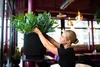 A woman is tending to a large potted plant in what appears to be a restaurant or cafe setting. Produktbild-Vorschau