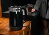 A black electric kettle is on a wooden counter, with a person holding a glass of dark liquid in the background. Podgląd zdjęcia produktu