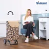 A woman is sitting on a chair reading a magazine next to a shopping trolley. Produktbild-Vorschau