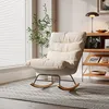 A cream-colored rocking chair with wooden rockers is placed on a patterned rug next to a window with white blinds and a tall green plant. A wooden shelf unit with books and a lamp is visible in the background. Náhľad obrázku produktu