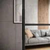 A modern living room interior with a grey sofa and wooden wall panels, viewed through a black framed glass door. Produktbild-Vorschau