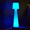 A tall, cone-shaped floor lamp with a pleated shade, illuminated with bright blue and cyan light, stands outdoors against a dark, slatted wall. Produktbild-Vorschau