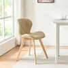A beige dining chair with wooden legs is placed on a white rug next to a white table, with a large window and curtains in the background. Produktbild-Vorschau