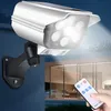 Weiße Solar-Sicherheitsleuchte mit mehreren LEDs, montiert an einer dunkelblauen Wand, hell leuchtend, mit einer Hand, die eine Fernbedienung im Vordergrund hält Voir les images du produit