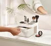 A hand is shown opening a drawer of a makeup organizer on a white dresser, with a mirror and a plant in the background. Náhled obrázku produktu