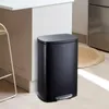 A black pedal trash can is shown in a kitchen setting next to a cabinet and a round rug. Produktbild-Vorschau