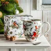 A Christmas mug and its packaging are displayed on a white surface next to a decorated Christmas tree. Voir les images du produit