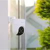 A white security camera mounted on a white post, with green foliage in the background. Produktbild-Vorschau