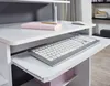 A close-up view of a white computer desk with a pull-out keyboard tray and a pink notebook. Produktbild-Vorschau